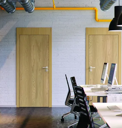 Dwa drewniane drzwi w biurze z białą ceglaną ścianą i nowoczesnymi biurkami oraz komputerami.