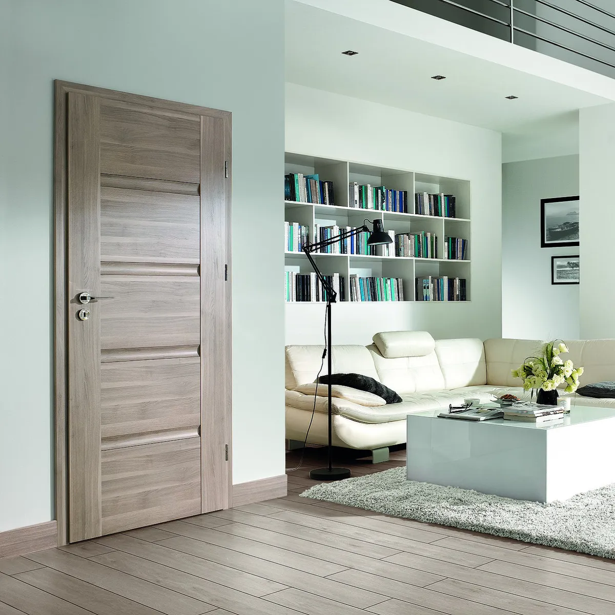 Nowoczesny salon z jasnymi ścianami, drewnianymi drzwiami, białą sofą i regałem z książkami.
