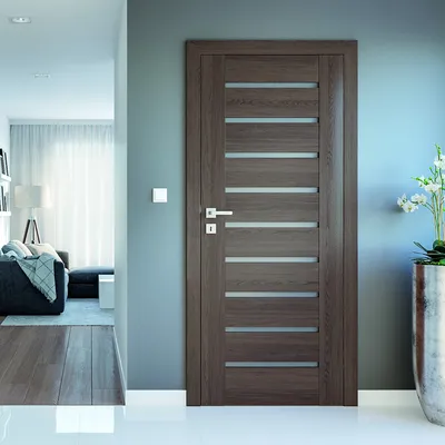 Nowoczesne drzwi w odcieniu drewna, prowadzące do jasnego pomieszczenia z kanapą i lampą. Obok kwiaty w doniczce.